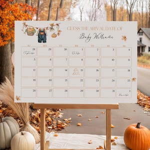 May include: A wooden easel displays a beige calendar with the text "GUESS THE ARIVAL DATE OF Baby Willene." The calendar is decorated with fall-themed illustrations. Pumpkins and fall foliage are in the foreground.