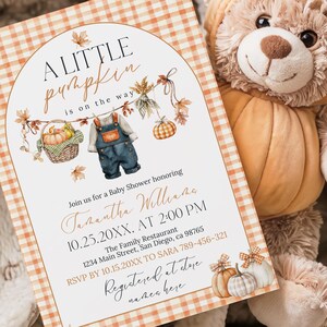 Puede incluir: Una invitaci&oacute;n a un baby shower con tem&aacute;tica de calabaza. La invitaci&oacute;n presenta el texto "A Little Pumpkin is on the Way" y los detalles del evento. Un oso de peluche con disfraz de calabaza est&aacute; junto a la invitaci&oacute;n sobre una manta suave.