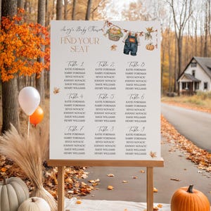 May include: A wooden easel displays a seating chart for a baby shower, with names listed under table numbers. The design features fall-themed illustrations of pumpkins and overalls. The scene is set outdoors with pumpkins and fall foliage.