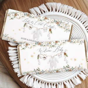 May include: Two rectangular baby shower favors with text that reads "Baby it's Cold Outside" and "Welcome Baby". The favors are decorated with a winter floral design and a baby onesie graphic. They rest on a white macrame coaster and a wooden tray.