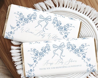 Puede incluir: Dos envoltorios de barra de chocolate blanco con diseños florales azules y letras doradas. El envoltorio superior dice "Bienvenido bebé William". El envoltorio inferior dice "Baby Shower de Avery. ¡Gracias por darnos tanto amor!"