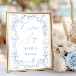 Op de afbeelding: Een ingelijst bord met een gouden rand, met een lichtblauw bloemmotief en de woorden "Custom Sign" en "TEXT HERE". Het bord bevat illustraties van een luchtballon, een teddybeer en een schommelpaard. Een boeket witte en lichtblauwe rozen staat op de achtergrond.