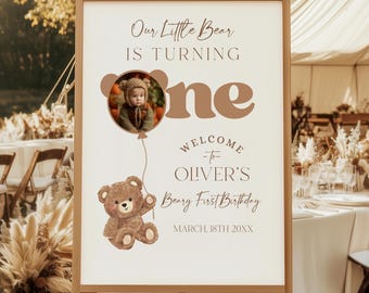 May include: A wooden easel displaying a welcome sign for a first birthday party. The sign features the text "Our Little Bear is Turning One" with a photo of a baby and a teddy bear illustration. The event is on March 18th, 20XX.