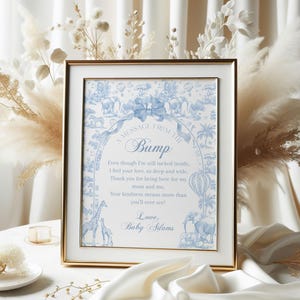 Op de afbeelding: Een ingelijste print met een gouden lijst, met een blauw en wit ontwerp met de tekst "A MESSAGE FROM THE Bump". De print bevat illustraties van dieren en luchtballonnen. Het frame staat op een witte tafel met een witte doek.