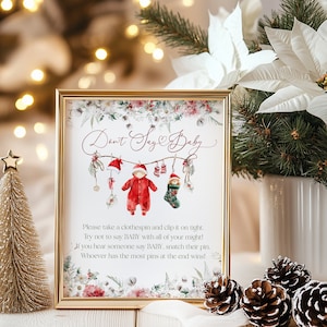 Puede incluir: Un letrero enmarcado con el texto "Don't Say Baby" e instrucciones para un juego de baby shower. El letrero presenta ilustraciones de ropa de bebé y decoraciones navideñas. También se ven un pequeño árbol de Navidad dorado y piñas.