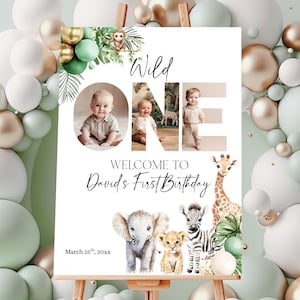 Può includere: Un cavalletto di legno espone un cartello di compleanno "Wild One" con foto di un bambino. Il cartello presenta illustrazioni di animali della giungla, il testo "Welcome to David's First Birthday" e la data 25 marzo 20xx. Lo sfondo include palloncini bianchi, dorati e verdi.