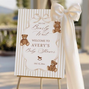 May include: A baby shower welcome sign on a gold easel. The sign has a cream and beige striped background with a decorative border. The sign reads "We Can Bearly Wait Welcome to Avery's Baby Shower" with teddy bear illustrations.