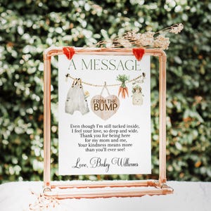 May include: A copper-colored metal frame holds a white sign with the text "A MESSAGE FROM THE BUMP." The sign features watercolor illustrations of baby clothes, carrots, and a bunny. The bottom of the sign reads "Love, Baby Williams."