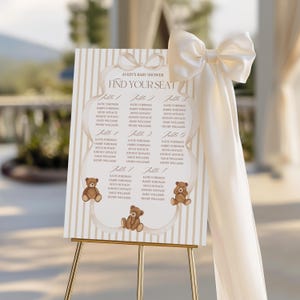 May include: A baby shower seating chart on a gold easel. The sign has a cream background with vertical stripes and a decorative border. The text "Avery's Baby Shower Find Your Seat" is at the top. Teddy bear illustrations are at the bottom.