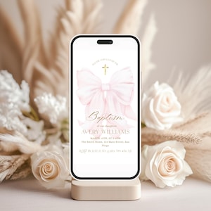 May include: A smartphone displays a baptism invitation. The design features a pink bow, a gold cross, and elegant script. The invitation includes the name "Avery Williams" and event details. The phone rests on a neutral-toned stand, surrounded by white roses and dried flowers.