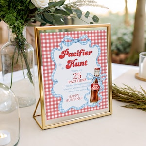 May include: A gold framed sign with the text "Pacifier Hunt" in red script. The sign has a red and white checkered background and a blue ribbon border. A bottle of soda is pictured on the sign. The sign is for a baby shower.