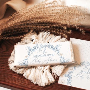 May include: A white chocolate bar with blue floral designs and the text "Amanda & David Baby Shower". The bar is on a wooden tray with a white tassel rug and a white card with blue floral designs and the text "Welcome Baby Williams".