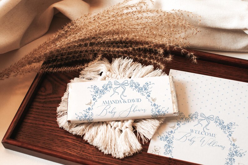 May include: A white chocolate bar with blue floral designs and the text "Amanda & David Baby Shower". The bar is on a wooden tray with a white tassel rug and a white card with blue floral designs and the text "Welcome Baby Williams".