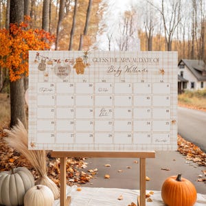 May include: A baby shower calendar with a plaid background and fall-themed decorations. The calendar reads "Guess the Arrival Date of Baby Williams." The calendar is displayed on a wooden easel, with pumpkins and fall foliage in the foreground.