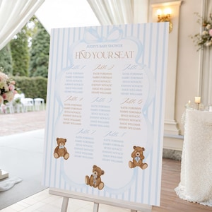 Puede incluir: Un plano de asientos para un baby shower sobre un caballete. El letrero tiene un fondo a rayas azules y blancas con un diseño de lazo azul claro en la parte superior. El texto "Avery's Baby Shower Find Your Seat" es de color cobre. Ilustraciones de osos de peluche marrones en la parte inferior.