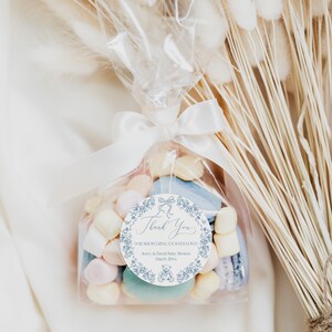 May include: A white favor bag filled with pastel-colored candies and macarons, tied with a white satin ribbon and a blue and white thank you tag that reads "Thank You for showering us with love! Avery & David Baby Shower March 20XX".