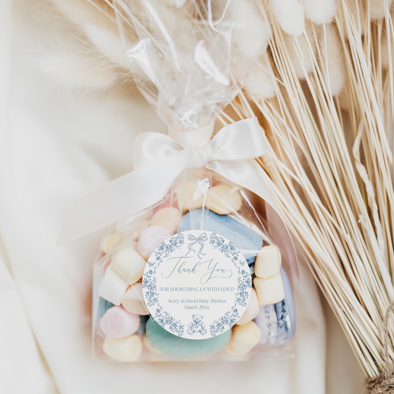 May include: A white favor bag filled with pastel-colored candies and macarons, tied with a white satin ribbon and a blue and white thank you tag that reads "Thank You for showering us with love! Avery & David Baby Shower March 20XX".