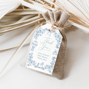 May include: A white thank you tag with blue floral designs and illustrations of a hot air balloon, a rocking horse, and a teddy bear. The text on the tag reads "Thank You Avery & David Baby Shower March 20XX". The tag is tied to a small brown burlap bag.