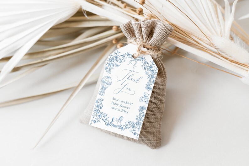 May include: A white thank you tag with blue floral designs and illustrations of a hot air balloon, a rocking horse, and a teddy bear. The text on the tag reads "Thank You Avery & David Baby Shower March 20XX". The tag is tied to a small brown burlap bag.
