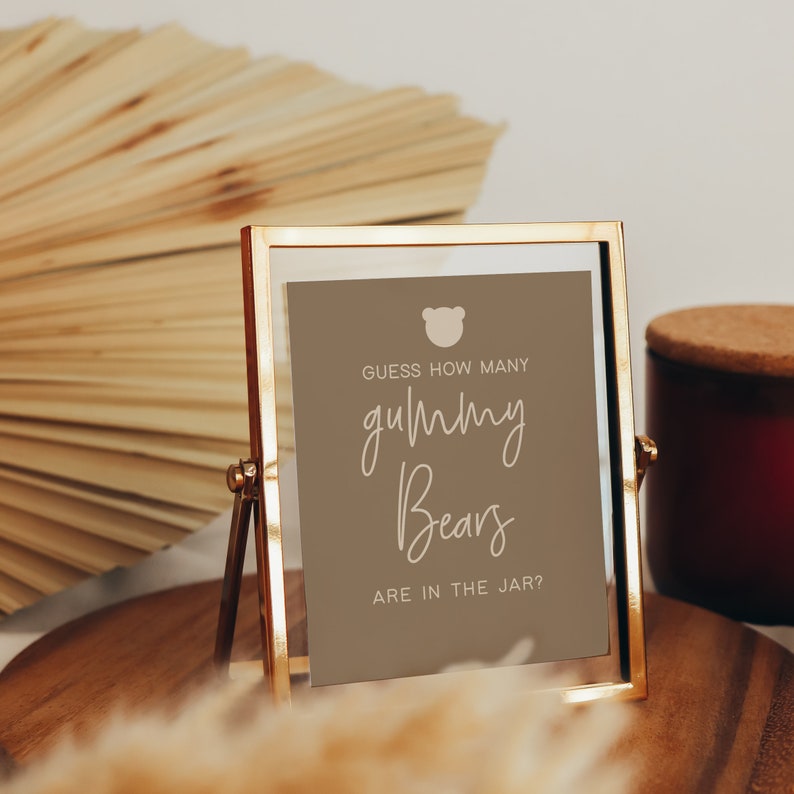 May include: A gold-framed sign with a brown background and white text that reads "Guess how many gummy bears are in the jar?" with a small bear icon in the top left corner.