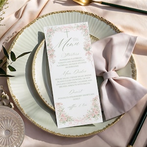 May include: A formal table setting with a menu card, plate, and silverware. The menu card features floral accents and the word "Menu" at the top. The plate is white with a gold rim, and the silverware has black and gold handles.