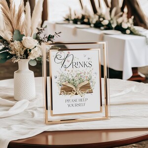 May include: A gold-framed sign with the word "Drinks" in elegant script, adorned with floral and butterfly illustrations. The sign reads "Please Help Yourself" and is set on a table with a white tablecloth, suggesting a wedding or event.