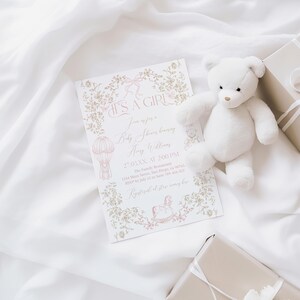 Op de afbeelding: Een uitnodiging voor een babyshower met de tekst "IT'S A GIRL" is omgeven door bloemmotieven en een illustratie van een luchtballon. Een witte teddybeer en twee ingepakte cadeaus met witte linten zijn ook aanwezig. De items zijn gerangschikt op een witte stof.