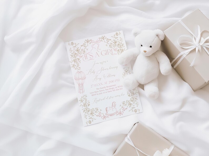 Op de afbeelding: Een uitnodiging voor een babyshower met de tekst "IT'S A GIRL" is omgeven door bloemmotieven en een illustratie van een luchtballon. Een witte teddybeer en twee ingepakte cadeaus met witte linten zijn ook aanwezig. De items zijn gerangschikt op een witte stof.
