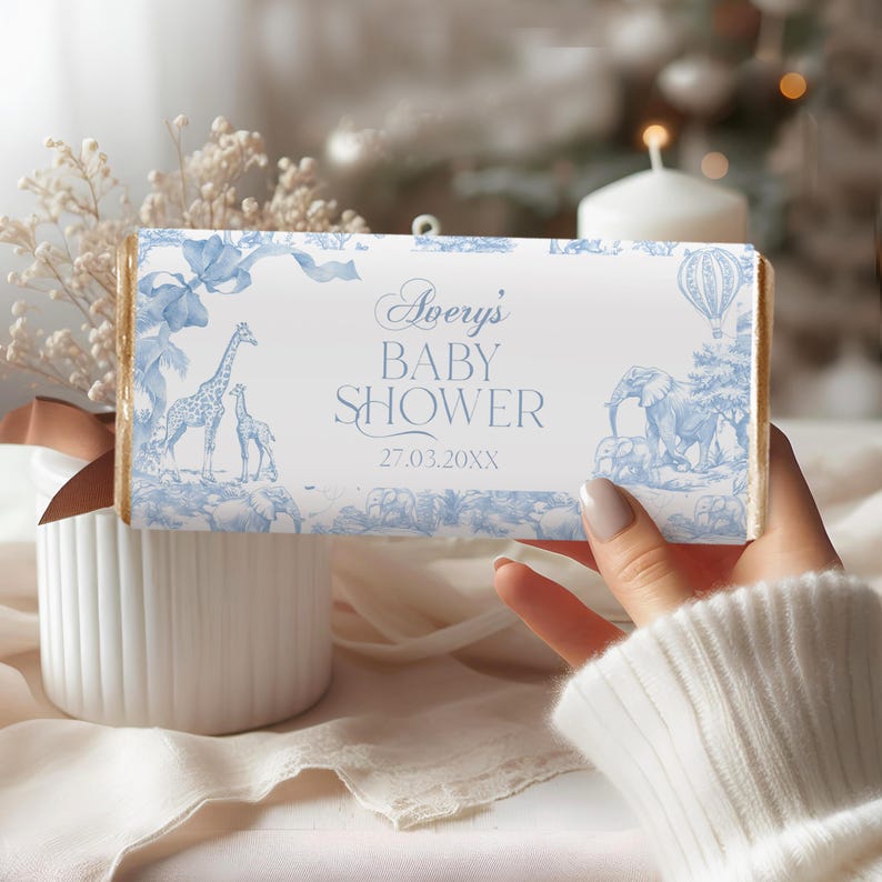 May include: A white chocolate bar wrapper with a blue and white design featuring a giraffe, elephant, and hot air balloon. The text on the wrapper reads "Avery's Baby Shower 27.03.20XX".