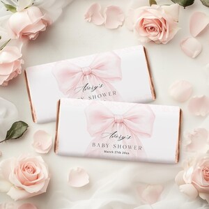 Könnte beinhalten: Zwei weiße Schokoladenriegelverpackungen mit einem rosa Schleifendesign und dem Text "Avery's BABY SHOWER". Die Verpackungen haben einen roségoldenen Rand und sind von rosa Rosen und Blütenblättern umgeben.