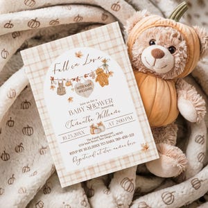May include: A baby shower invitation with a fall theme. The invitation features the text "Fall in Love" and "Baby Shower" with decorative elements like leaves and teddy bears. A plush teddy bear in a pumpkin costume rests beside the invitation.