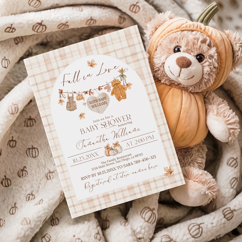 May include: A baby shower invitation with a fall theme. The invitation features the text "Fall in Love" and "Baby Shower" with decorative elements like leaves and teddy bears. A plush teddy bear in a pumpkin costume rests beside the invitation.