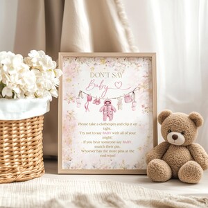 Puede incluir: Un letrero enmarcado para el juego de baby shower con el texto "DON'T SAY Baby" y un gráfico de tendedero. Un oso de peluche marrón está sentado a la derecha. Una cesta de mimbre con flores blancas está a la izquierda.