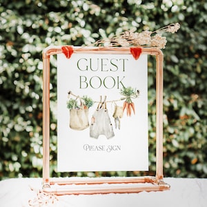 May include: A copper-colored metal frame holds a white sign with the words "GUEST BOOK" in green script. Below, watercolor illustrations depict a tote bag, overalls, and carrots hanging on a clothesline. The sign reads "PLEASE SIGN" in a decorative font.