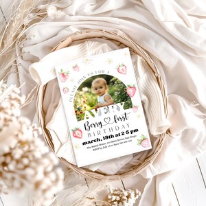 May include: A white invitation card with a photo of a baby, strawberries, and daisies. The text reads "Avery Berry first Birthday" and includes the date and time. The card is in a woven basket.