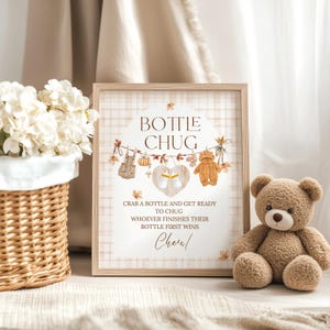 May include: A framed sign with the text "Bottle Chug" and a teddy bear illustration. The sign is decorated with baby-themed graphics and the text "Crab a bottle and get ready to chug. Whoever finishes their bottle first wins." A brown teddy bear sits beside the sign.