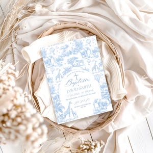 May include: A baptism invitation card with a blue and white safari animal design. The card includes the text "Baptism of David Smith" and event details. The invitation is placed in a woven basket with a white fabric background.
