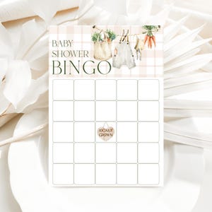 Może przedstawiać: Drukowana karta do gry w bingo na baby shower z r&oacute;żowo-białym tłem w kratkę. Karta ma tytuł "Baby Shower Bingo" i przedstawia szereg ilustracji ubranek dla dzieci i warzyw wiszących na sznurku. Centralne pole ma znak w kształcie serca z napisem "Lokalnie uprawiane".