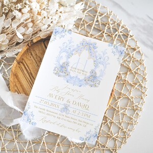 Op de afbeelding: Een blauwe en gouden babyborrel uitnodiging met een koets ontwerp en bloemmotieven. De tekst luidt "Baby Shower Honoring Avery & David 27.05.20XX van 14:00-15:00 uur The Family Restaurant 1234 Main Street, San Diego, CA 98765. RSVP voor 15 juli naar 789-456-321 Geregistreerd bij [naam van de winkel]"