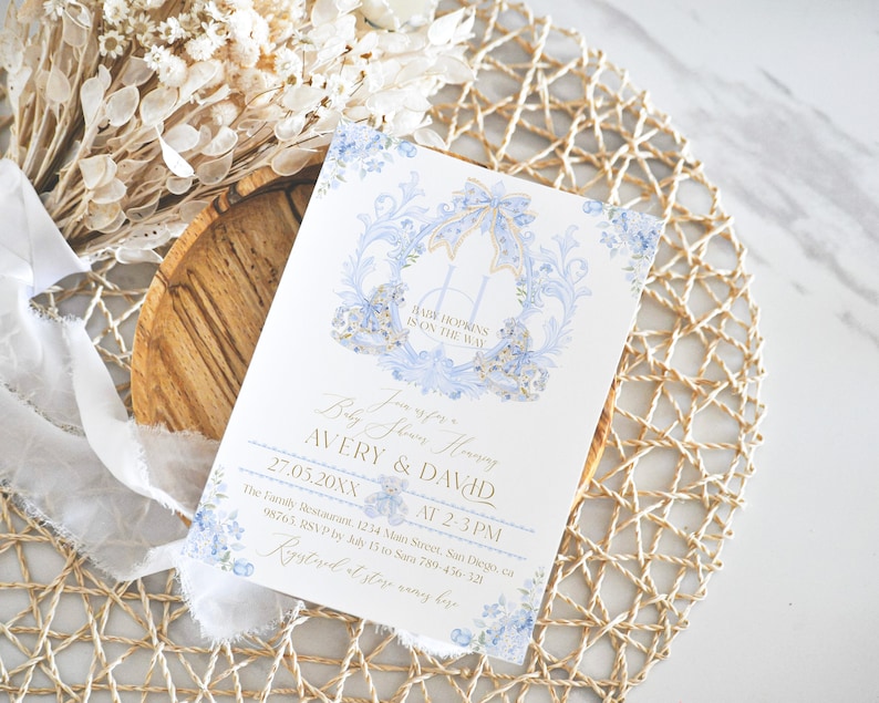 Op de afbeelding: Een blauwe en gouden babyborrel uitnodiging met een koets ontwerp en bloemmotieven. De tekst luidt "Baby Shower Honoring Avery & David 27.05.20XX van 14:00-15:00 uur The Family Restaurant 1234 Main Street, San Diego, CA 98765. RSVP voor 15 juli naar 789-456-321 Geregistreerd bij [naam van de winkel]"