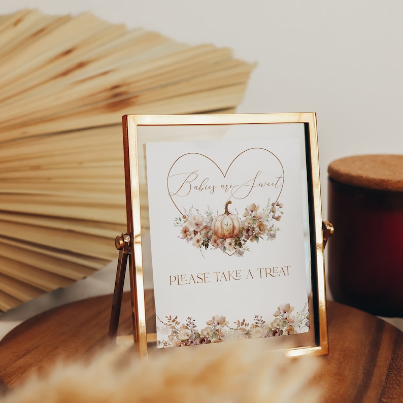 Pu&ograve; includere: Un cartello con cornice dorata con la scritta "Babies are Sweet" all'interno di una forma a cuore, con un design a zucca e floreale. Il cartello dice anche "PLEASE TAKE A TREAT". Il cartello &egrave; su una superficie di legno.