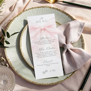May include: A formal table setting with a menu card. The menu card features a pink bow design and a list of menu items. The plate is white with a gold rim, and a pink napkin is folded in a bow shape. Gold utensils complete the setting.