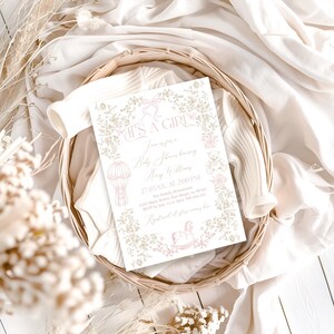 Op de afbeelding: Een uitnodiging voor een babyshower met de tekst "IT'S A GIRL" in elegant schrift, omringd door bloemmotieven en decoratieve elementen. De uitnodiging heeft een zachtroze en beige kleurenpalet, met illustraties van een luchtballon, teddybeer en schommelpaard. De uitnodiging is geplaatst in een gevlochten mand.