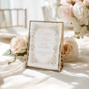 May include: A gold framed sign with the text "Custom Sign" and "Fully Editable" surrounded by a floral border of pink roses and green leaves. The sign is on a white surface with roses in the background.