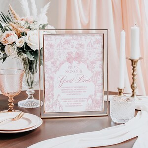 May include: A framed pink guest book sign with the text "PLEASE SIGN OUR Guest Book". The sign has a pink and white pattern with giraffes and other animals. A bouquet of flowers, a glass, and candles are also visible.