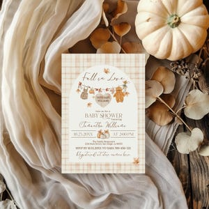 May include: A baby shower invitation with a fall theme. The invitation features the text "Fall in Love" and "Baby Shower" with a heart and teddy bear graphic. The background has a plaid pattern and is decorated with pumpkins and leaves.