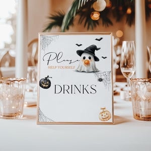 May include: A framed Halloween-themed sign with a gold border, featuring a cartoon ghost wearing a witch hat, bats, and the text "Please Help Yourself DRINKS." Two candle holders and wine glasses are on the table.