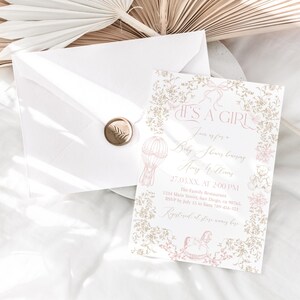 Op de afbeelding: Een uitnodiging voor een babyshower met de tekst "IT'S A GIRL" in een sierlijk handschrift. De uitnodiging heeft een roze en gouden bloemenontwerp, een illustratie van een luchtballon en een schommelpaard. Een envelop met een gouden zegel is ook aanwezig.