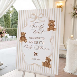Puede incluir: Un letrero de baby shower sobre un caballete con un fondo a rayas crema y beige. El letrero dice "We can bearly wait" y da la bienvenida a los invitados a "Avery's Baby Shower". Se presentan ilustraciones de osos de peluche marrones.