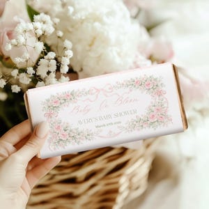 May include: A rectangular baby shower favor with a pink and gold design. The favor has the text "Baby In Bloom" and "Avery's Baby Shower March 27th 2025" in a decorative floral frame with pink roses and bows.
